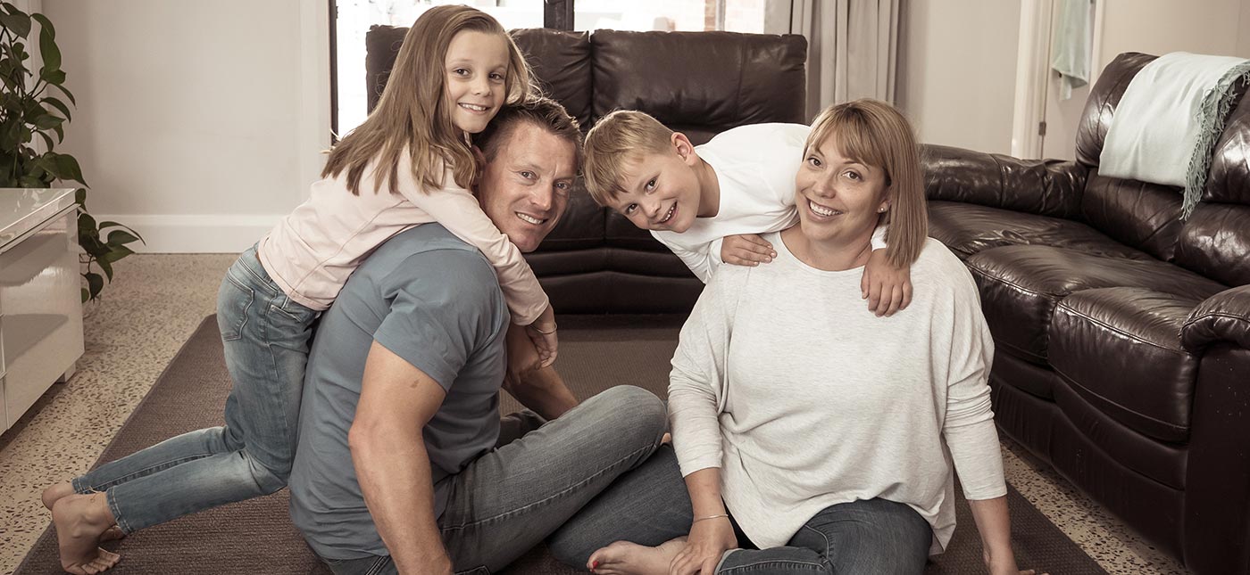 A family sitting on the floor of their living room (One guy, one girl and two kids), a sofa set just around them